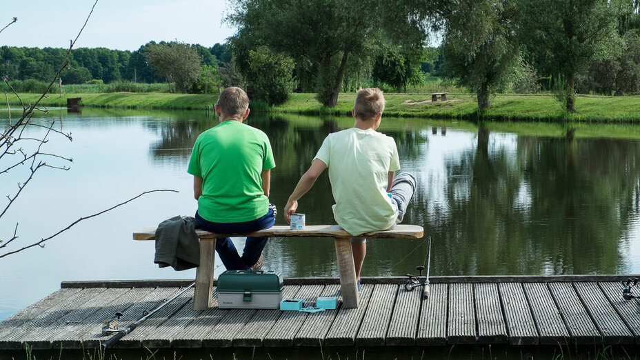 Zwei Männer sitzen mit Angelzubehör auf einer Bank am See.