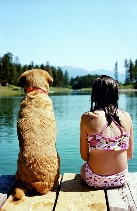 Auf einem Holzsteg am See sitzen ein brauner Hund und ein Mädchen nebeneinander mit dem Rücken zur Kamera. 