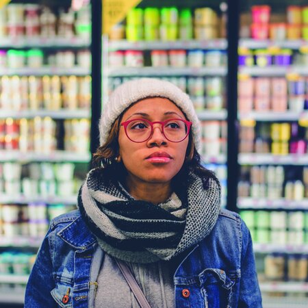 Eine junge Frau mit Mütze und Brille steht in einem Supermarkt vor dem Kühlregal.